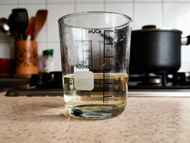Verre doseur contenant liquide jaune, posé sur comptoir de cuisine; poêle visible en arrière-plan.
