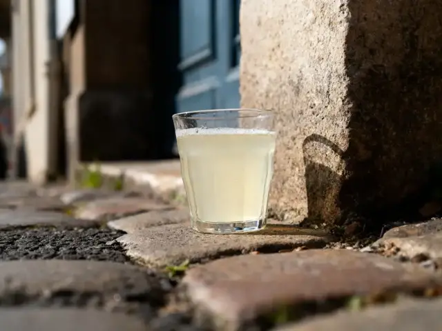 Verre de boisson jaune pâle sur pavés, ombre projetée sur mur ensoleillé.