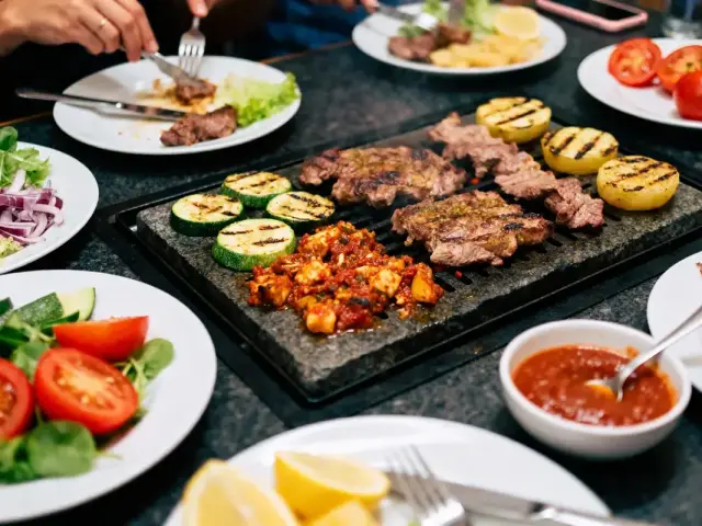 Assiette de grillades, légumes et sauce sur table partagée. Mains avec fourchettes, ambiance conviviale.
