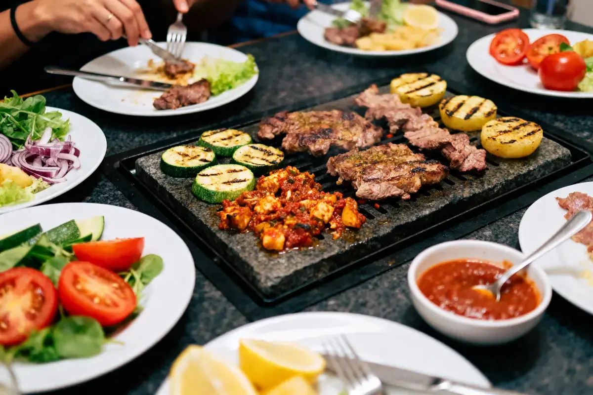 Assiette de grillades, légumes et sauce sur table partagée. Mains avec fourchettes, ambiance conviviale.