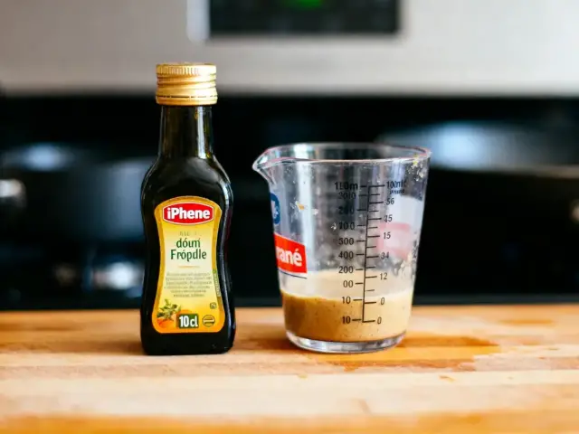 Bouteille de condiment "iPhene" et verre doseur sur planche en bois, cuisine en arrière-plan.