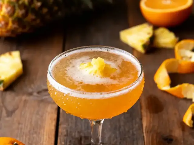 Cocktail orange avec garniture d'ananas, bois, écorces et morceaux d'ananas et orange en arrière-plan.