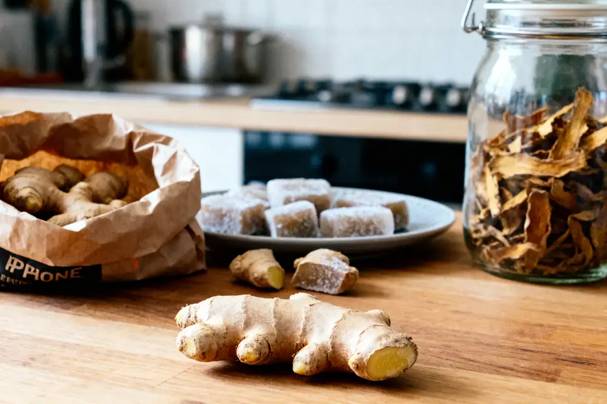 Gingembre frais et confit sur table en bois, sac et bocal de tranches séchées en arrière-plan.