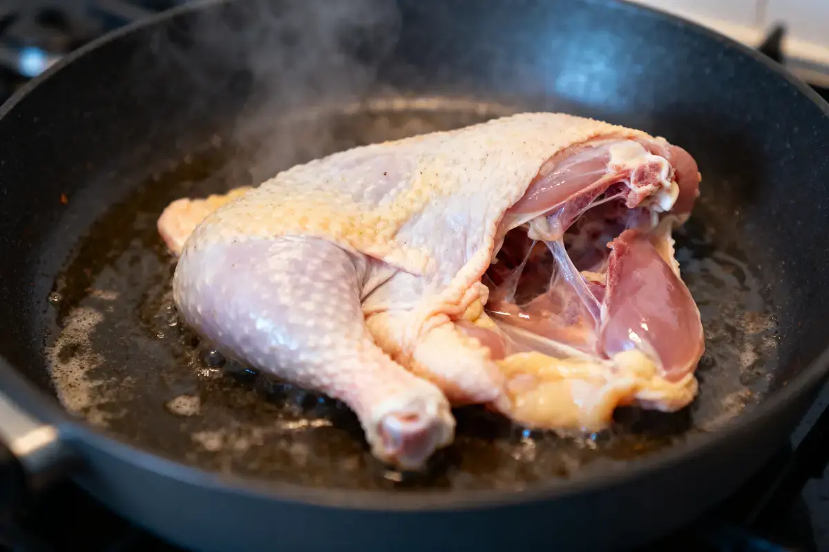 Poulet entier dans une poêle sur feu, en cours de cuisson, dégagement de vapeur visible.