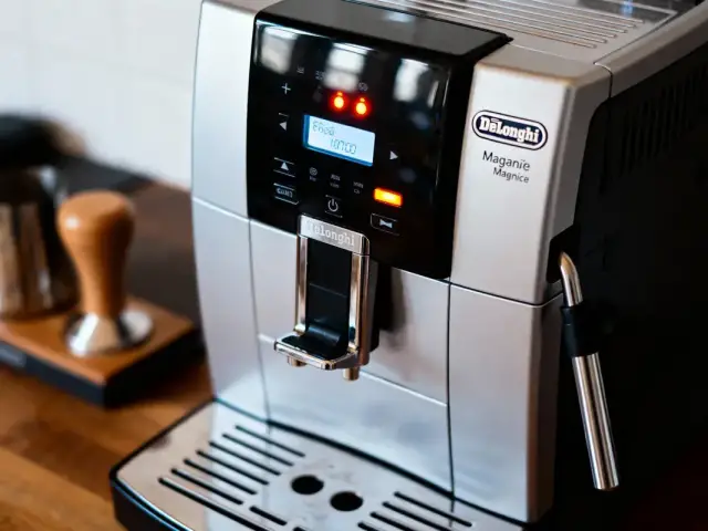 Machine à café argentée DeLonghi avec écran numérique allumé, tamper et mousseur à lait en arrière-plan.