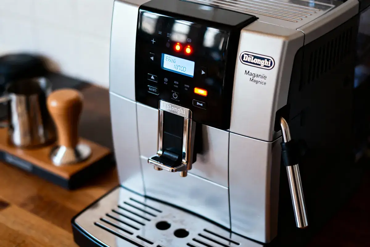 Machine à café argentée DeLonghi avec écran numérique allumé, tamper et mousseur à lait en arrière-plan.