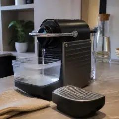 Machine à café sur plan de travail, carafe en verre, corbeille de fruits, essuie-tout beige.