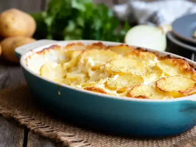 Gratin dauphinois dans un plat bleu sur une table en bois, pommes de terre en arrière-plan.