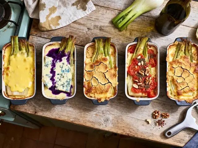 Quatre plats de gratin de fenouil variés sur une table en bois, avec bouteille d'huile et économe.