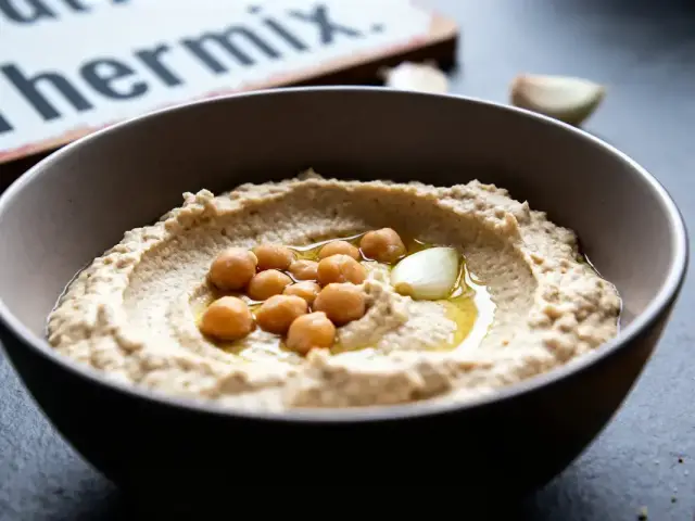 Bol de houmous garni de pois chiches et huile d'olive, avec pain en arrière-plan.