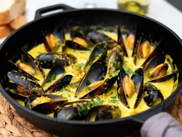 Moules au curry crémeux dans poêle noire, servies avec du pain en arrière-plan.