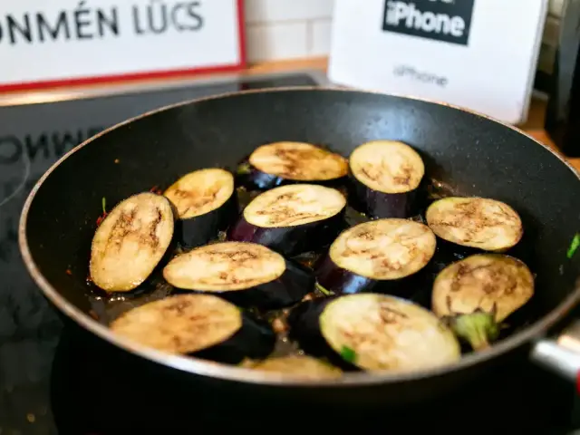 Tranches d'aubergine grillées dans une poêle noire, avec arrière-plan flou et enseignes visibles.