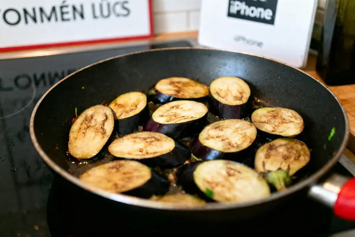 Tranches d'aubergine grillées dans une poêle noire, avec arrière-plan flou et enseignes visibles.