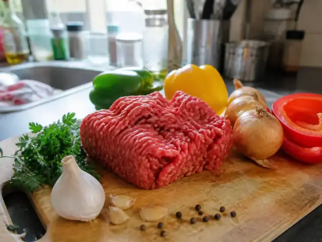 Viande hachée avec poivrons, oignons, ail, persil sur planche en bois dans une cuisine lumineuse.