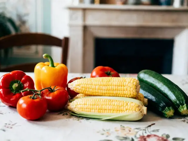 Légumes variés sur une table : poivrons, tomates, maïs, courgettes, sur nappe fleurie devant cheminée.