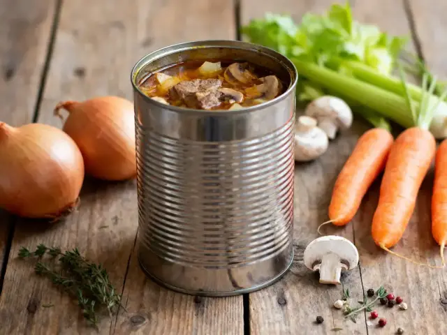 Boîte de conserve avec ragoût, carottes, oignons, céleri, champignons, épices sur table en bois rustique.