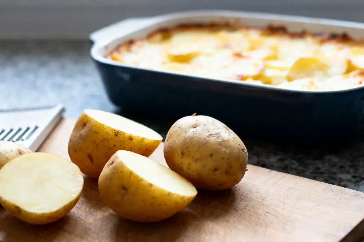 Pommes de terre coupées sur planche, gratin de pommes de terre au four en arrière-plan.