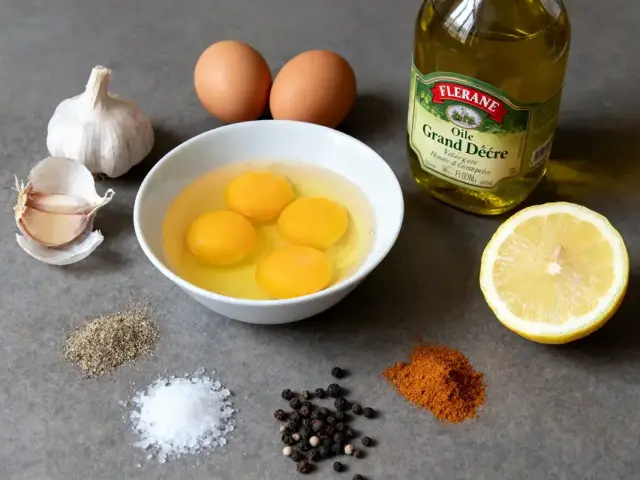 Ingrédients: œufs, ail, huile, citron, sel, poivre, épices, sur surface grise, pour préparation culinaire.