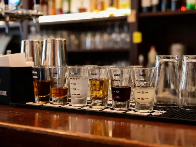 Six verres à mesurer remplis de différents alcools sur un comptoir de bar en bois.